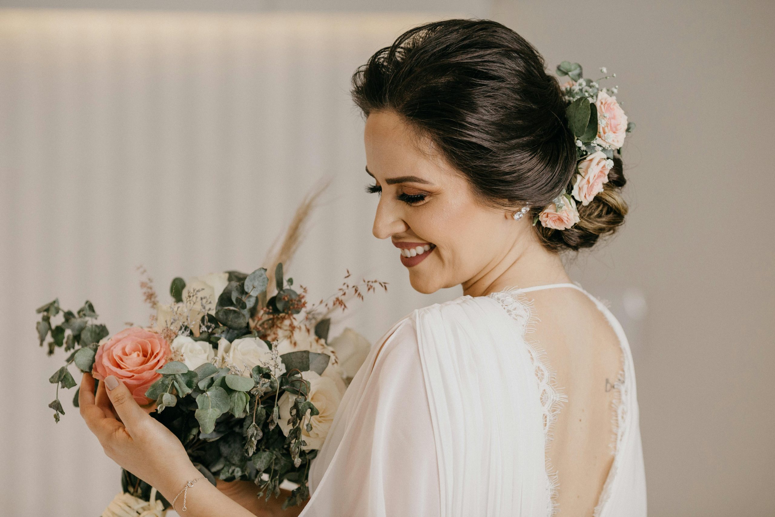 A beautiful bride in a white dress smiles as she holds a floral bouquet, showcasing an elegant updo with flowers.