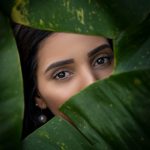 A woman's eyes gaze through lush green leaves, creating a mysterious and natural portrait.